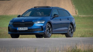 Blauer Skoda Superb Combi schräg von vorn vor Wiese aufgenommen. Schiebedach ist geöffnet.