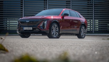 Cadillac Lyriq schraeg von vorn fotografiert vor modernem Gebäude, Cadillac ist in rotmetallic lackiert