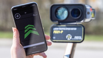 ayern, München: Eine Hand hält ein Smartphone mit einer Blitzerwarnapp, während im Hintergrund ein Radar-Messgerät zu sehen ist. (zu dpa: «Handy-Führerschein soll kommen - Diskussion um Blitzerapps») Foto: Lino Mirgeler/dpa/dpa-tmn +++ dpa-Bildfunk +++