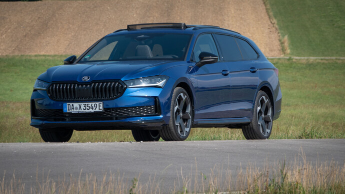 Blauer Skoda Superb Combi schräg von vorn vor Wiese aufgenommen. Schiebedach ist geöffnet.
