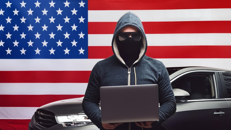 Fotorealistische Darstellung eines Hackers mit Sonnenbrille, Kapuze und Notebook. Er steht vor einem Auto. Im Hintergrund ist die US-Flagge sichtbar.